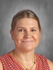 Headshot of a smiling woman with light brown hair, wearing a red patterned top, set against a neutral background.
