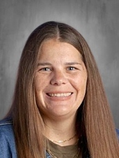 A smiling woman with long brown hair, wearing a denim jacket and a green shirt, against a neutral background.