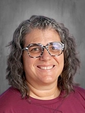 Smiling woman with curly gray hair and glasses, wearing a maroon shirt, set against a neutral background.