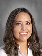 Smiling woman with long brown hair, wearing a white blouse, against a gray background.