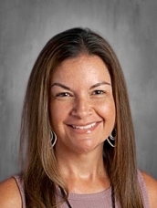 Headshot of a woman with long brown hair, wearing a sleeveless top and earrings, smiling against a neutral background.