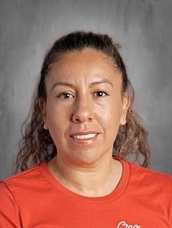 Portrait of a woman with brown skin and curly hair, wearing an orange shirt, smiling against a gray background.