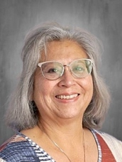 Smiling woman with gray hair and glasses, wearing a colorful patterned top, against a neutral background.