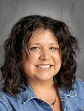 Smiling woman with curly dark hair, wearing a denim jacket, against a neutral background.