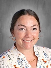 A woman with brown hair, wearing a floral blouse, smiles against a gray background.