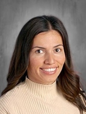 A smiling woman with long brown hair, wearing a beige turtleneck, poses against a gray backdrop.