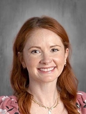 Smiling woman with long red hair, wearing a floral top and necklace, set against a neutral background.