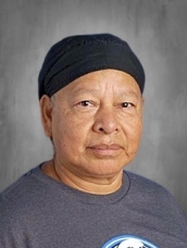 An older person with a serious expression, wearing a black headband and a gray t-shirt, against a gray background.