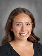 Smiling young woman with long brown hair, wearing a black top, against a gray background.