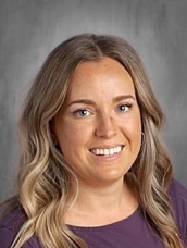 A smiling woman with long, wavy hair and wearing a purple shirt, posed against a neutral background.