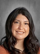 Portrait of a woman with long dark hair, smiling, wearing an orange sweater against a neutral background.