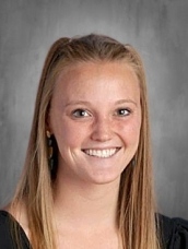 Smiling young woman with long brown hair, wearing a black top, against a gray background.