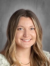 A smiling woman with long, wavy hair, wearing a light-colored top, poses for a professional headshot.