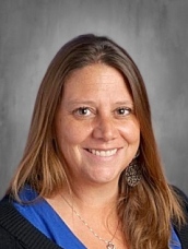 Smiling woman with long brown hair, wearing a blue shirt and earrings, against a gray background.