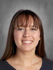 A smiling woman with long brown hair and bangs, wearing a dark shirt, against a neutral background.