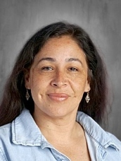 A woman with long dark hair and earrings smiles at the camera, wearing a light blue denim jacket.