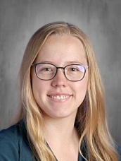 A smiling person with long blonde hair and glasses, wearing a dark top, against a gray background.