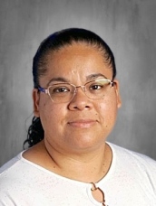 A woman with glasses and a ponytail smiles at the camera, wearing a white shirt against a gray background.