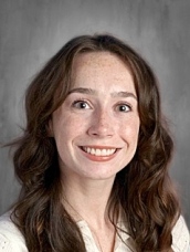 Smiling young woman with long, wavy brown hair and freckles, wearing a light-colored shirt, against a gray background.