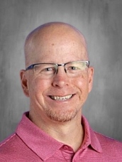 Smiling man with glasses and a bald head, wearing a pink polo shirt, against a gray background.