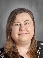 Portrait of a woman with long brown hair, smiling, wearing a black top with decorative patterns, against a gray background.