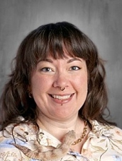 Smiling woman with long brown hair and a floral shirt, wearing facial piercings, against a gray background.