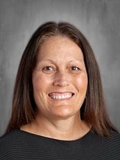 Headshot of a woman with long brown hair, smiling, wearing a black shirt, with a neutral gray background.