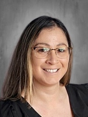 A smiling woman with long brown hair and glasses, wearing a black top, against a gray background.