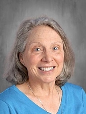 Smiling woman with gray hair, wearing a blue shirt, against a neutral background.
