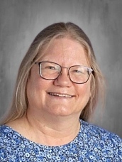 Smiling woman with long, light brown hair and glasses, wearing a blue floral top, against a gray background.