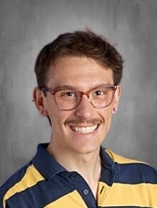 Smiling person with short hair, glasses, and a mustache, wearing a blue and yellow striped polo shirt.