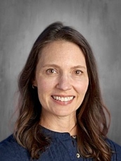 A smiling woman with long brown hair, wearing a dark blue top, against a neutral background.
