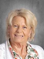 Smiling older woman with blonde hair, wearing a floral blouse and a white cardigan, against a plain background.