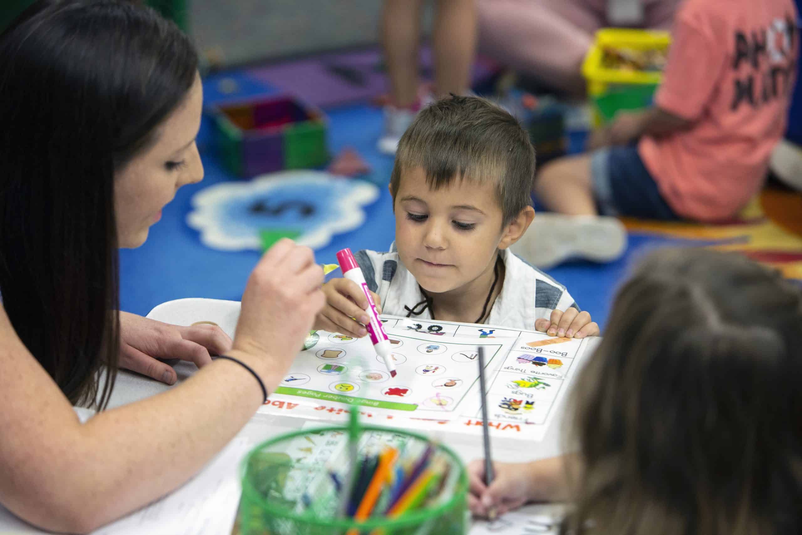 Preschool boy writing in classroom with help by female teacher.