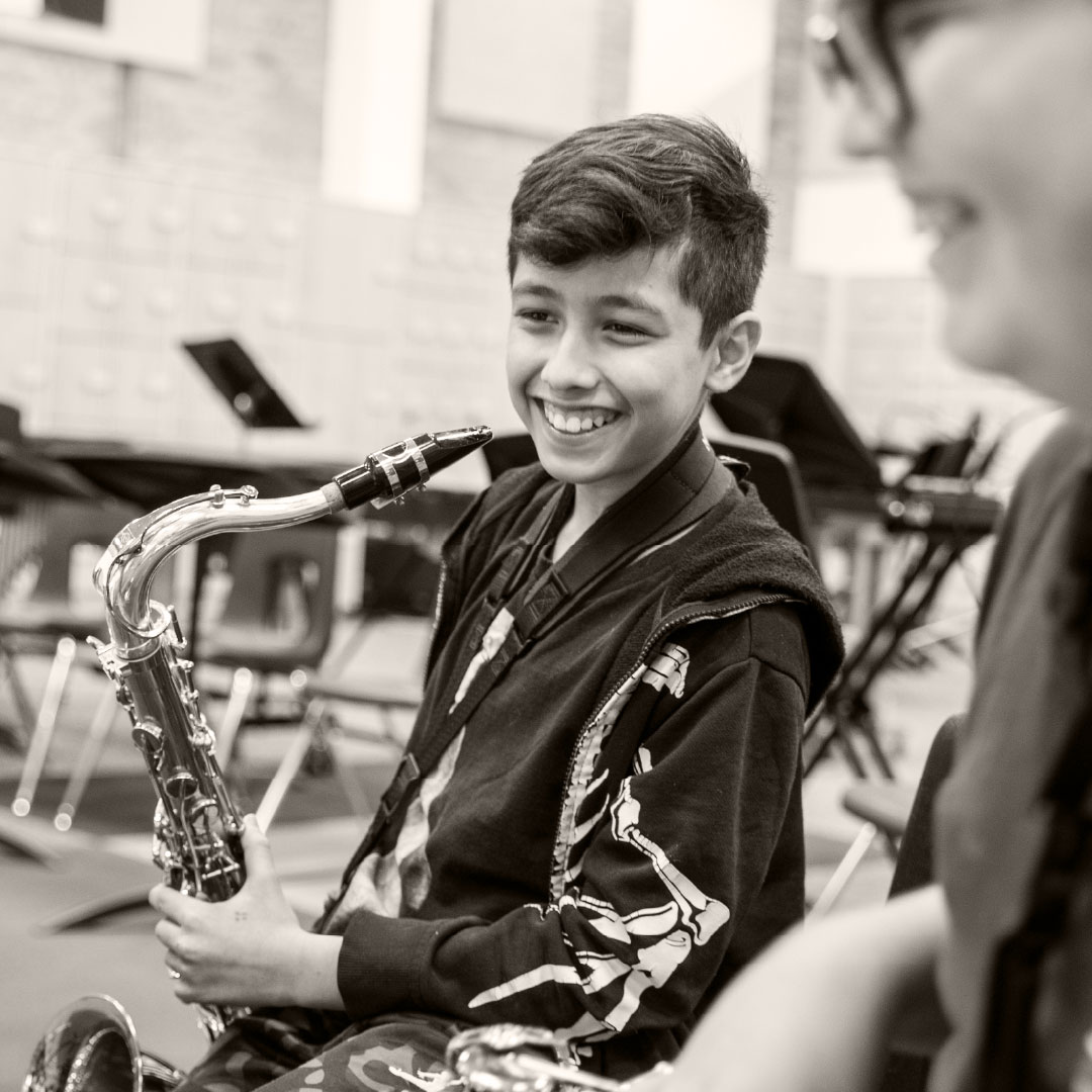 A smiling boy holds a saxophone in a music classroom, with a blurred classmate nearby.