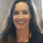 A woman with long dark hair smiles at the camera, wearing a black top and a beaded necklace, with a textured wall behind her.