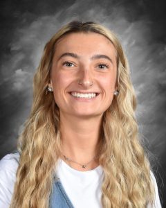 Smiling person with long blonde hair, wearing a white top and denim vest, against a gray background.