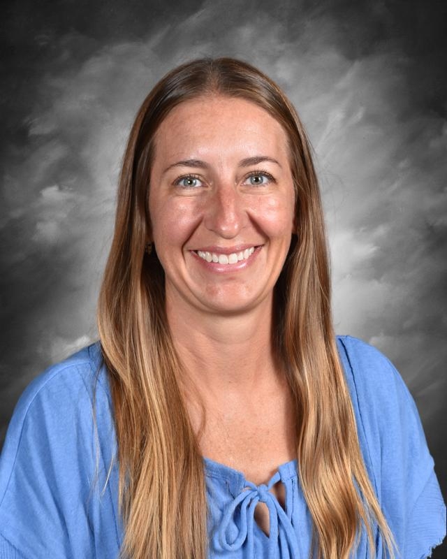 A woman with long hair smiles at the camera, wearing a blue blouse against a gray background.