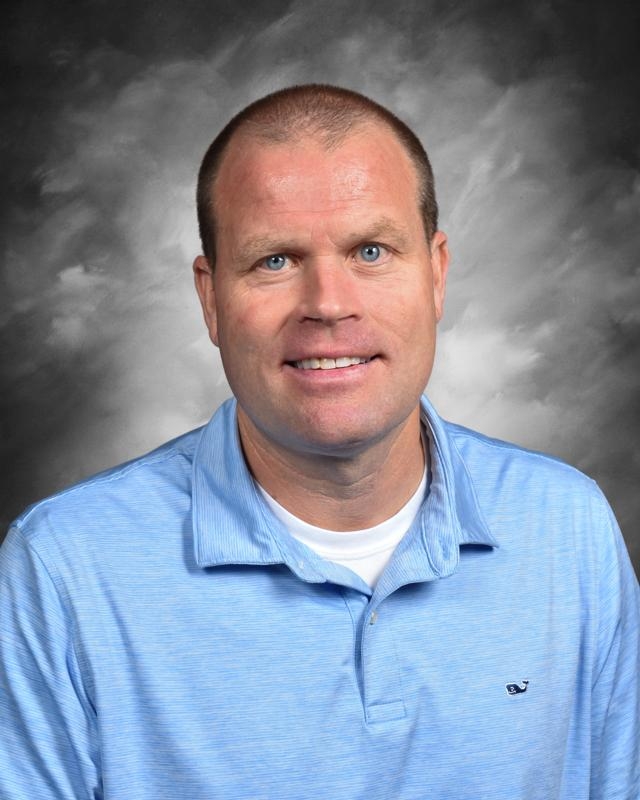 A man in a light blue polo shirt smiles against a gray, cloudy background.