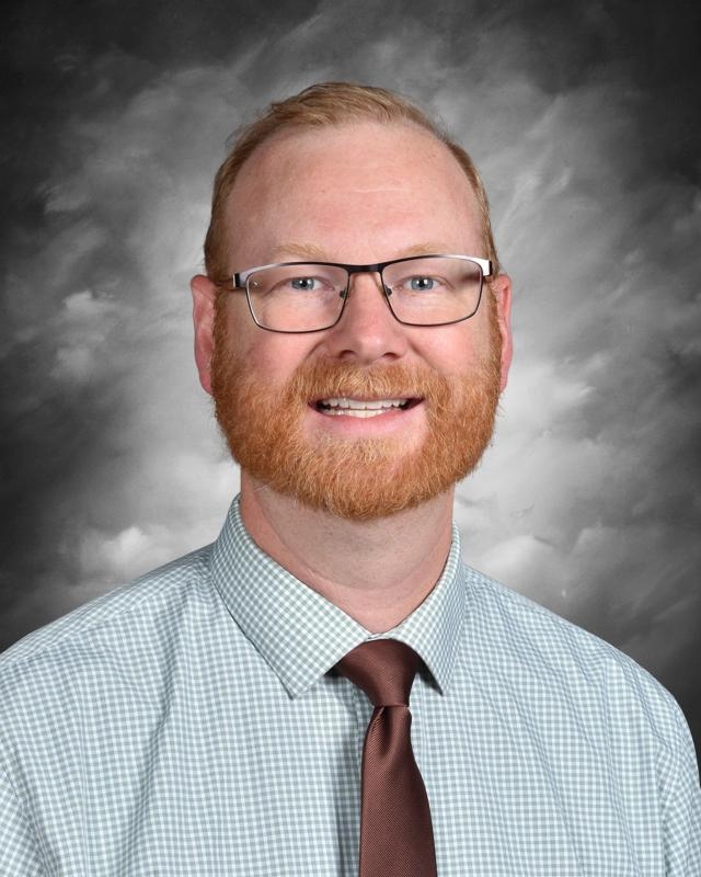 A person with glasses, a beard, and a tie smiles in front of a cloudy background.