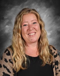 Smiling woman with long wavy hair, wearing a patterned cardigan over a black top, against a gray background.