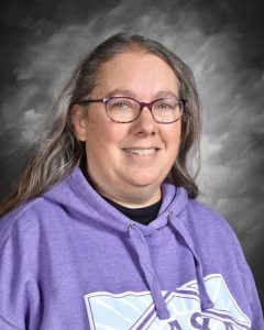 Smiling person with long hair and glasses, wearing a purple hoodie, against a gray background.