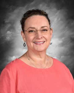 Smiling woman with glasses, wearing a coral top, against a gray cloudy background.