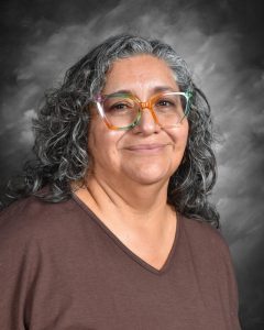 A person with curly hair and colorful glasses smiles against a gray background.